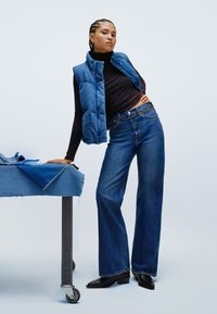 Blauwe puffer gilet over een zwarte coltrui, gecombineerd met hoog getailleerde blauwe flare jeans. Het model staat naast een tafel met denimstof.