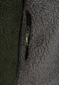Gros plan d'une veste polaire vert foncé et grise avec une fermeture éclair noire et une languette en tissu vert portant l'inscription « UNFR ».