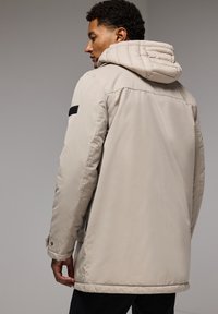 Chaqueta beige aislante con capucha, que presenta una tela texturizada, bolsillos laterales y un detalle negro en el hombro.