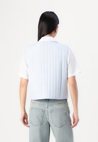 Kobieta z długimi czarnymi włosami, ubrana w jasnoniebieską pikowaną kamizelkę na białą koszulę z krótkim rękawem i jasnoniebieskie dżinsy, odwrócona plecami.