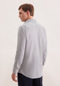Uomo girato di lato che indossa una camicia a maniche lunghe a quadretti piccoli e pantaloni scuri su uno sfondo chiaro e uniforme.