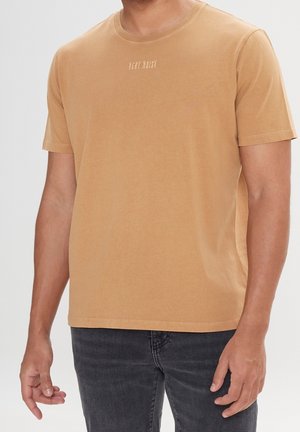 Man draagt een lichtbruine korte mouw t-shirt met de tekst "VERY NOISY" op de borst en donker gewassen spijkerbroek, staand tegen een lichtgrijze achtergrond.