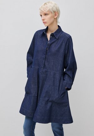 Osoba z krótkimi blond włosami ubrana w granatową sukienkę dżinsową z długim rękawem i dużymi kieszeniami, stojąca na tle jasnego, gładkiego tła.