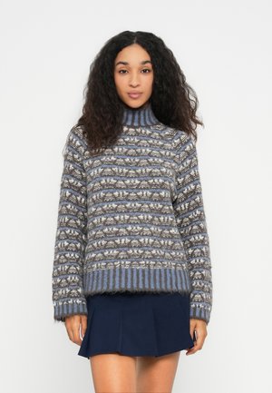 Frau mit langen, lockigen Haaren, die einen gemusterten Rollkragenpullover in Blau, Grau und Weiß sowie einen marineblauen, gefalteten Minirock trägt und vor einem weißen Hintergrund steht.