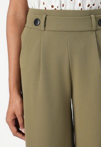 Pantaloni verde oliva con una texture liscia, caratterizzati da una vita alta, due bottoni neri e pieghe nette che esaltano l'aspetto sartoriale.