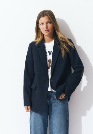 Blazer de cuadro marino con cierre de dos botones, solapas con muesca y parches en los codos. Combinado con una camiseta gráfica blanca y jeans holgados.