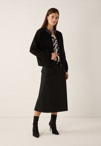 Schwarze Bomberjacke aus strukturiertem Stoff mit glänzendem Kragen und Reißverschlusstaschen. Kombiniert mit einem schwarzen Rock und Stiefeln mit hohen Absätzen.