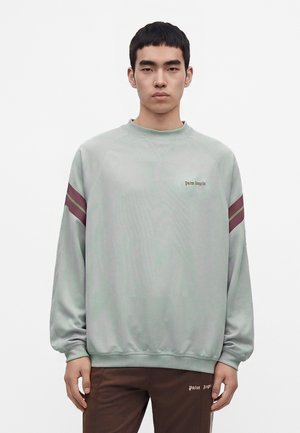 Jeune homme aux cheveux courts noirs, portant un sweat-shirt gris clair avec des rayures bordeaux aux manches et un pantalon marron, sur un fond uni.