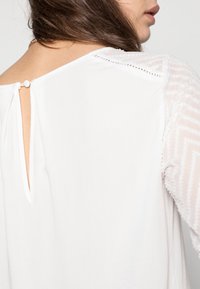 Blusa bianca con scollatura rotonda, presenta una chiusura sul retro a forma di chiave con un bottone e maniche trasparenti testurizzate con un motivo a zigzag.