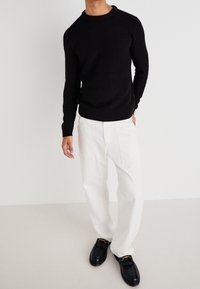 Czarny prążkowany sweter dziany, białe spodnie o prostych nogawkach, czarne mokasyny ze złotymi akcentami. Prosty, swobodny strój zaprezentowany na neutralnym tle.