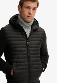 Schwarze Puffer-Weste mit Reißverschluss, strukturierter gepolsterter Vorderseite und glatten Ärmeln. Mit Kapuze und einem auffälligen Markenlogo auf der Brust.