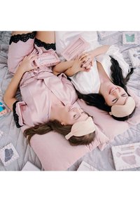 Jedwabna bielizna nocna w różowo-białych kolorach; akcenty z koronki; miękka różowa poduszka; użytkownicy trzymają się za ręce, nosząc jasne opaski na oczy; obok znajdują się kosmetyki.