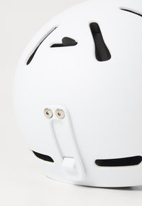 Hvit hjelm med ventileringsåpninger, justerbar dial og bakfeste. Glatt overflate med synlig maskinvare og en logo nær bunnen.