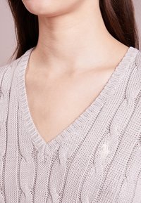 Pull gris clair en tricot avec un col en V, un motif torsadé et des bords côtelés. Le matériau semble doux et texturé.