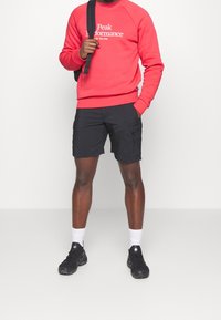 Rode sweatshirt met wit logo, zwarte cargoshorts met zijzakken, wit en zwart sokken, en zwarte sportschoenen.