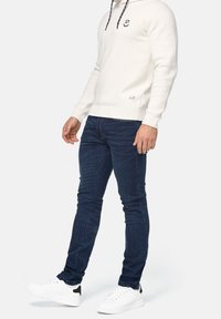 Lysefarvet hættetrøje med snøre i hætten, mørkeblå jeans og hvide sneakers med sort detalje på hælen. Minimalistisk logo på hættetrøjen.