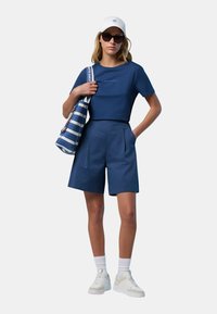 Maglietta blu navy con testo stampato, pantaloni corti blu navy abbinati, sneakers bianche, calze bianche e una borsa a righe, completata da un cappellino bianco.