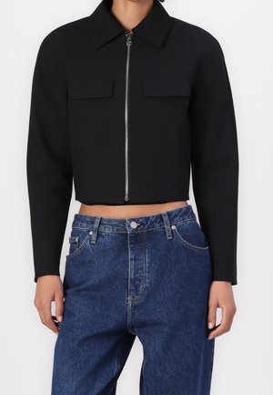 Person iført en sort cropped jakke med lynlås foran og to brystlommer, kombineret med højttaljede blå denimjeans.