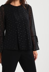 Blusa nera con maniche lunghe trasparenti, caratterizzata da una parte anteriore plissettata e accenti metallici sparsi. Lacci alle aperture delle maniche.