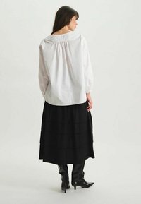 Chemise blanche à manches longues avec détail froncé dans le dos, associée à une jupe noire à volants et des bottes noires en cuir montantes.