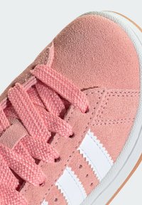 Light pink suede sneaker with textured surface, white leather accents, and woven pink laces. Features a flat white rubber sole.