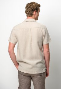 Camicia di lino beige a maniche corte con piega sul retro, colletto e orlo leggermente curvo. Abbinata a pantaloni corti color marrone chiaro, che mostrano la texture naturale del tessuto.