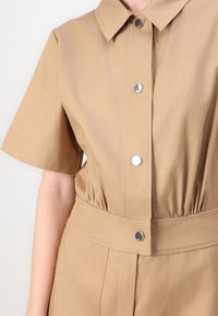 Väljä beige haalari, jossa on lyhyet hihat, napitettava etuosa ja vyötäröstä kiristetty malli. Sisältää metalliset napit ja sileän, kevyen kankaan tekstuurin.