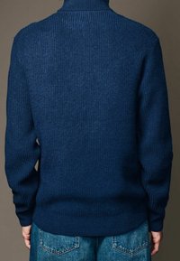 Pull en maille côtelée bleu marine avec un col montant, doté de longues manches et d'un ourlet ajusté. Porté avec un jean en denim bleu clair.