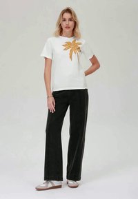 Balta trumpa rankovėmis marškinėliai su auksine palmės atspaudu, derinami su juodomis plačiomis kelnėmis su aukštu liemeniu ir šviesios spalvos bateliais.