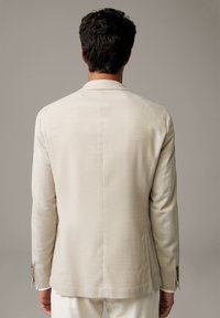 Uomo dai capelli scuri che indossa un blazer beige e pantaloni bianchi, girato di spalle contro uno sfondo grigio uniforme.