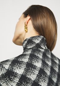 Czarna i biała koszula w kratę z wysokim kołnierzem. Złote teksturowane kolczyki. Gładka tkanina o usztywnionym fasonie, z nachodzącymi wzorami.