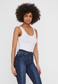 Débardeur blanc à encolure dégagée et bretelles fines, associé à un jean taille haute bleu foncé avec une texture lisse et un léger stretch.