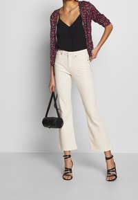 Mujer vestida con jeans acampanados color crema, tacones negros con tiras, camiseta negra de tirantes, chaqueta corta floral, sosteniendo un pequeño bolso negro.