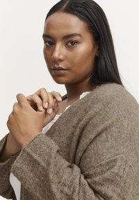 Knit sweater in a soft brown tone with textured fabric, v-neck, and loose sleeves. Model displays hands in a relaxed pose.
