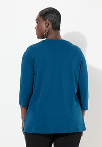 Top teal a maniche lunghe con scollo rotondo e spacco laterale, realizzato in tessuto morbido. Presenta una vestibilità rilassata e una texture liscia.
