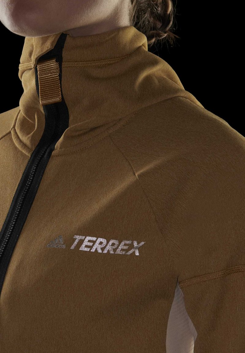 Brązowa kurtka zapinana na zamek z wysokim kołnierzem, z czarnym zamkiem błyskawicznym i odblaskowym, srebrnym logo "TERREX" na klatce piersiowej. Gładka faktura materiału.