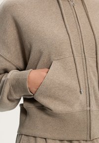 Felpa beige con zip realizzata in materiale morbido, caratterizzata da un cappuccio regolabile con coulisse, polsini a coste e una tasca frontale con una leggera texture.