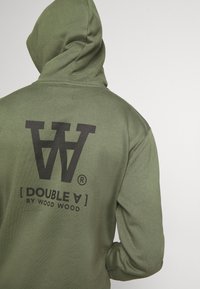 Sudadera verde oliva con un gran logo negro en la parte posterior, bolsillo estilo canguro en la parte delantera, tejido texturizado y capucha con cordón.