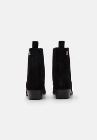 Botas de tobillo de ante negro con una textura suave, que cuentan con un tacón bloque, tiradores laterales y un detalle en oro en el tobillo exterior.