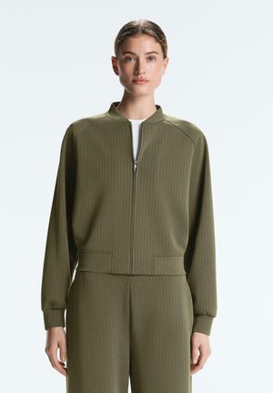 Žena oblečená v olivovo zelenej bunde na zips a zodpovedajúcich nohaviciach s jemnými vertikálnymi pruhmi, stojaca pred jednoduchým svetlým pozadím.