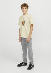 T-shirt gialla chiara con stampa grafica, maniche corte, abbinata a jeans di denim grigi e sneakers nere con dettagli bianchi.