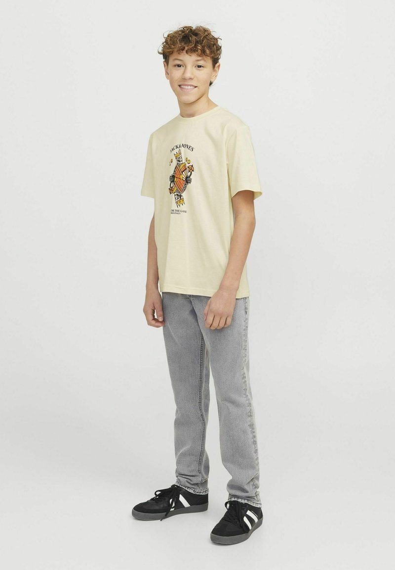 T-shirt gialla chiara con stampa grafica, maniche corte, abbinata a jeans di denim grigi e sneakers nere con dettagli bianchi.