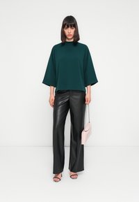 Top overdimensionato color teal con maniche corte, abbinato a pantaloni in finta pelle nera. Una elegante borsa rosa e sandali neri con lacci completano il look.