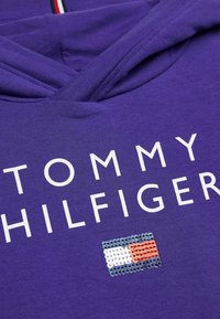 Fialová mikina s kapucí, na které je bílý nápis "TOMMY HILFIGER" a výšivka loga se sequin v modré, bílé a červené barvě. Měkká textilie.