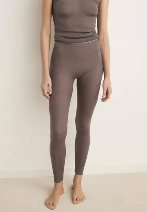 Person som bär en matchande taupe-ribbade linne och leggings, står barfota på ett ljust golv mot en enkel vägg.