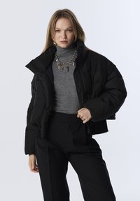 Veste doudoune noire courte avec finition brillante, dotée d'un col montant et de boutonnières. Associée à un col roulé gris et un pantalon noir.