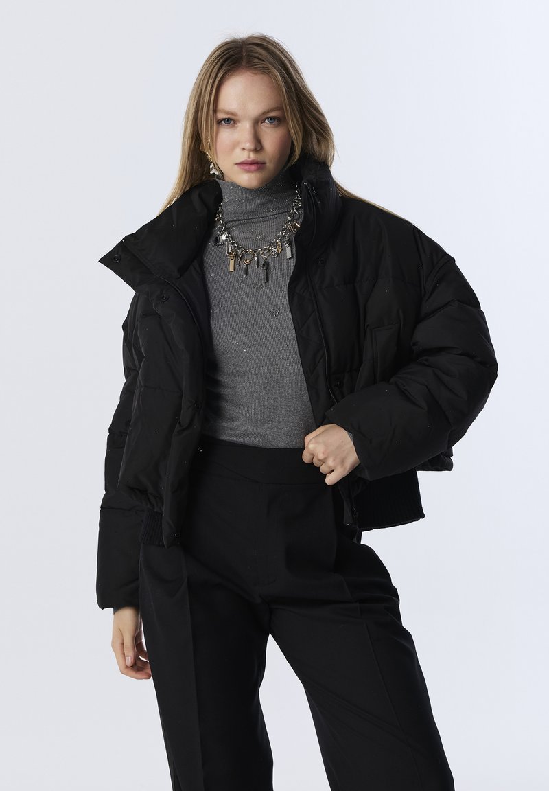 Veste doudoune noire courte avec finition brillante, dotée d'un col montant et de boutonnières. Associée à un col roulé gris et un pantalon noir.