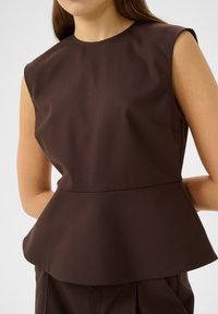 Smeđa bezrukavna peplum majica od teksturiranog materijala s okruglim izrezom i suptilnom bočnom džepom. Čist, strukturiran dizajn.