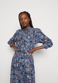 Tamsiai mėlynos spalvos ilgomis rankovėmis blusas, puošta įvairių spalvų gėlių raštu. Ji turi apykaklę ir mygtukų užsegimą priekyje.