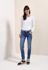 Witte top met lange mouwen gecombineerd met blauwe jeans met een slanke pasvorm en zichtbare details van het voorvak. Donkere sneakers maken de outfit compleet.